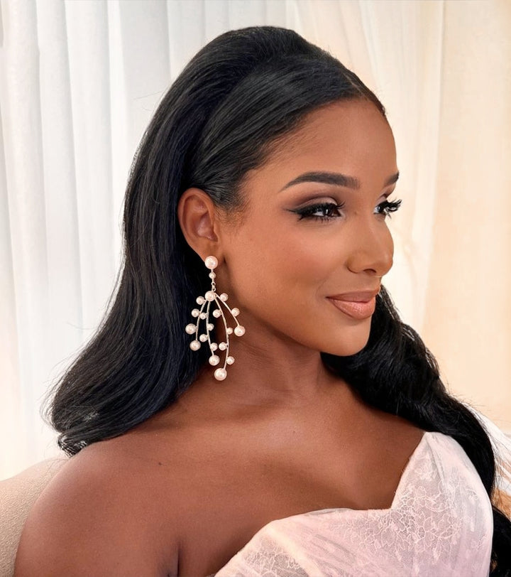 A woman with long, wavy black hair and glamorous makeup wears Modern Pearl Chandelier Earrings (style no. E216) with a soft pink off-shoulder dress, posing in front of pale curtains.