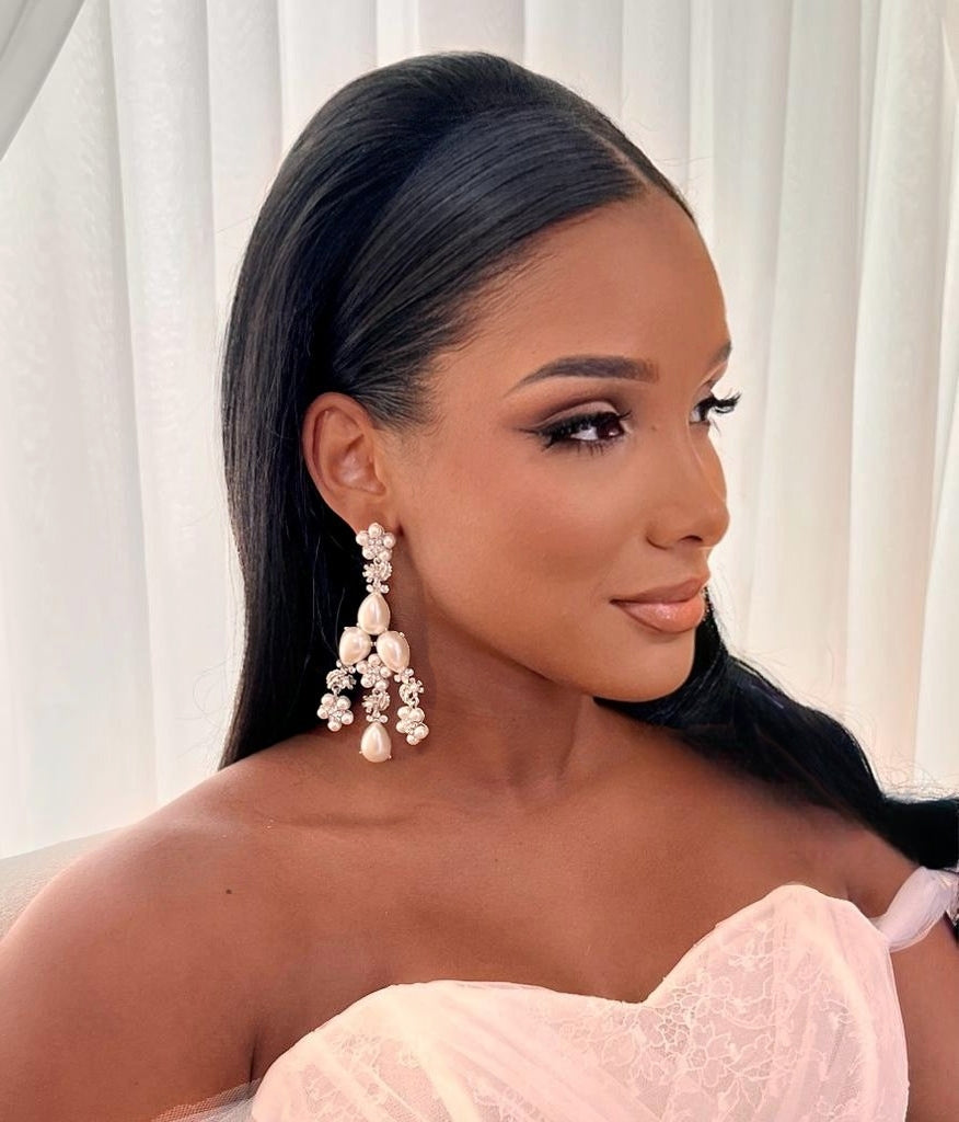 A woman with long black hair wears a light pink off-shoulder dress and Floral Pearl Statement Chandelier Earrings (style no. E215). Sitting before white curtains, she gazes serenely to the side.
