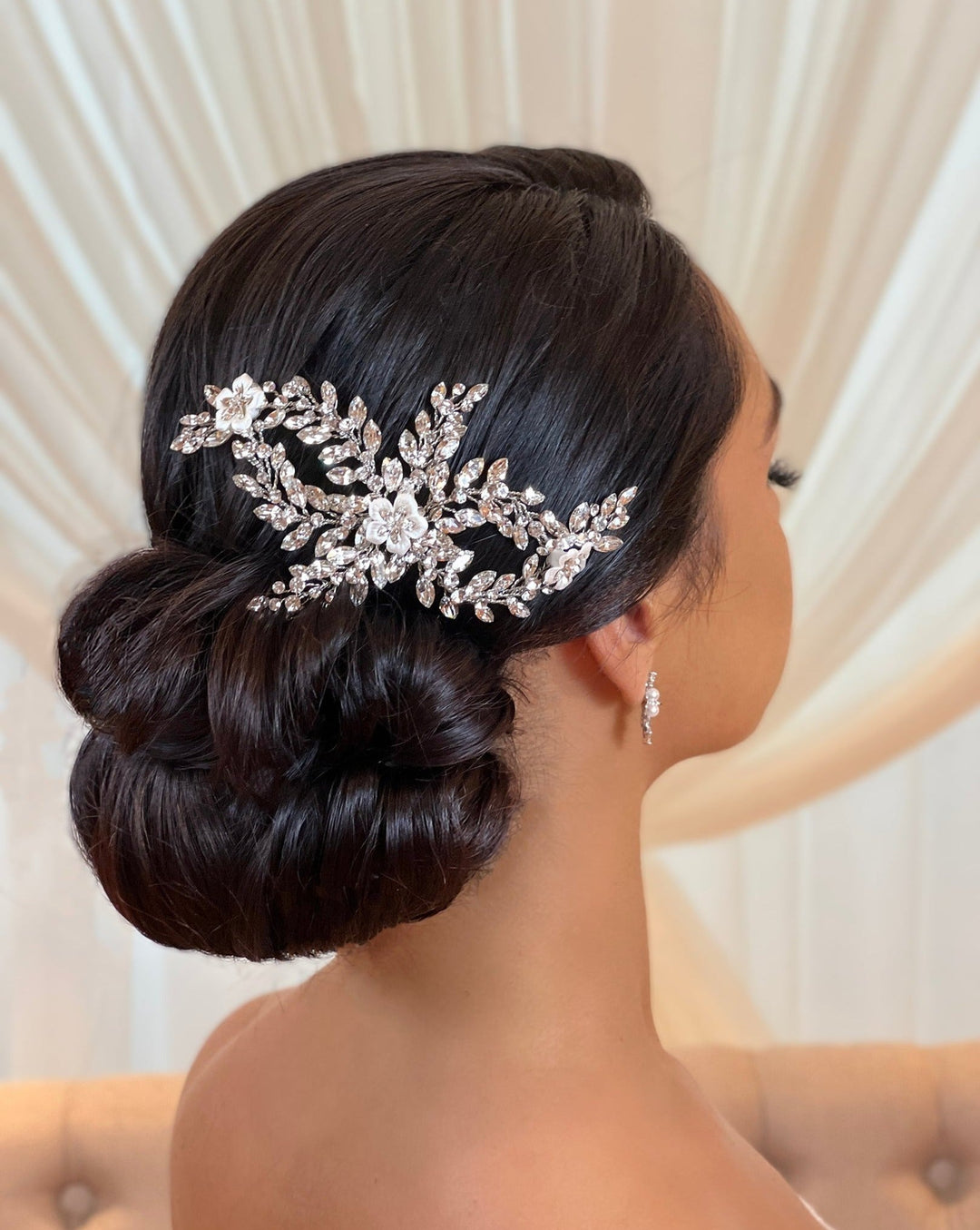 A woman with dark hair styled in an elegant low bun is adorned with the Swarovski Crystal and Porcelain Flower Bridal Comb (style no. 242726) and small hoop earrings, set against a softly blurred background of light-colored draped fabric.