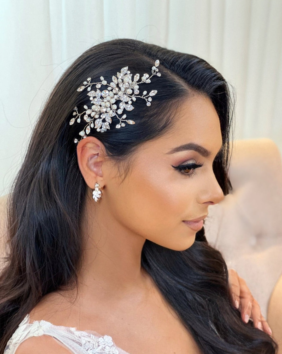 A woman with long black hair is seated, adorned with the Delicate Pearl and Swarovski Crystal Hair Comb (style no. 242691). She wears matching CZ wedding earrings and a white outfit. Her makeup features elegant, defined eyeliner, set against a soft, neutral background.
