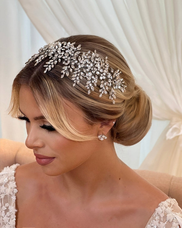 A woman with blonde hair styled in an elegant updo wears a sparkling, ornate Glistening Cluster Swarovski Crystal Hair Vine style no. 3951567 with leaf designs. She has subtle makeup and Cubic Zirconia wedding earrings.