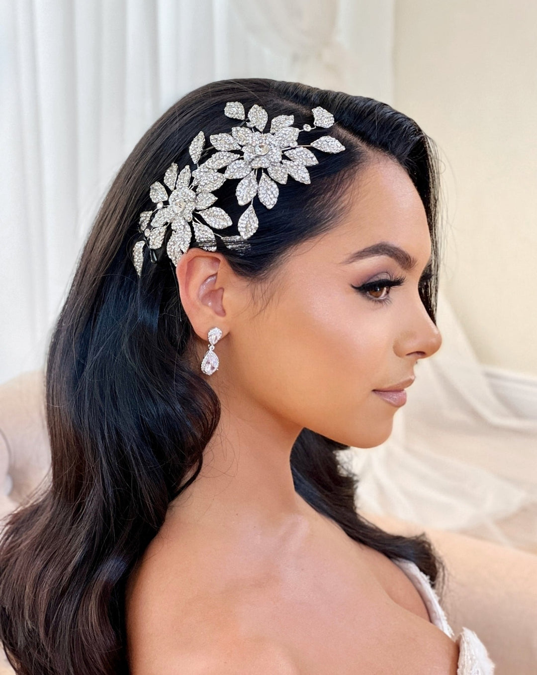 A woman with long dark hair is shown in profile, wearing the sparkling Dazzling Double Blossom Bridal Hair Comb (style no. 80065) and drop earrings. Her elegant, subtle makeup complements the soft, neutral indoor setting.