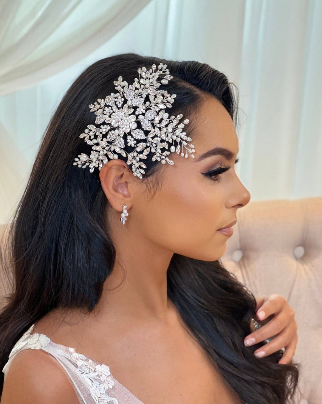 A woman with long dark hair wears a Floral Crystal and Pearl Hair Comb (style no. 242716) paired with matching CZ wedding earrings. Shes seated in profile on a beige upholstered chair against sheer white curtains framing the background.