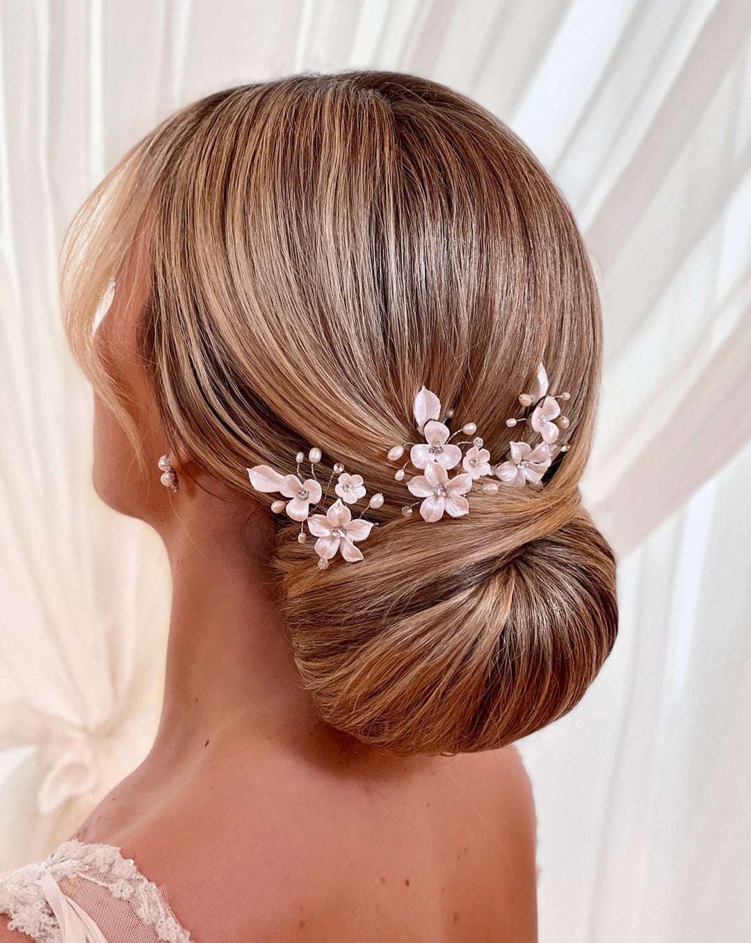 A person with light brown hair styled in an elegant, low bun adorned with a Porcelain Flower Pin (style no. 242723) featuring Swarovski crystals, delicate white floral designs, and pearl accents against sheer white curtains.