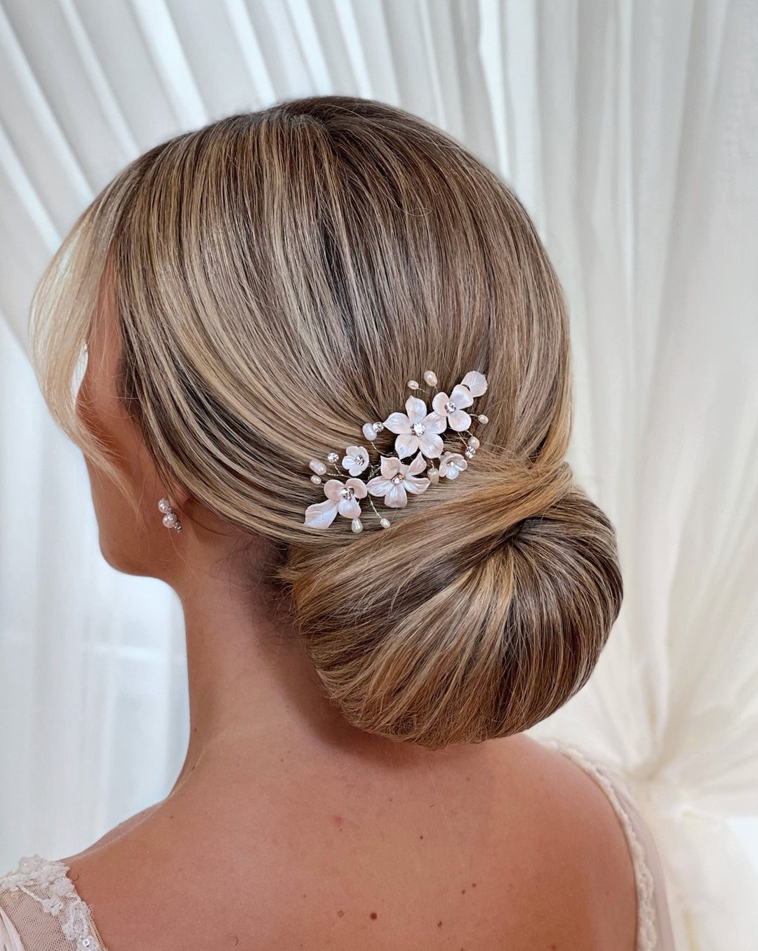 A woman with light brown hair styled in a low bun wears the Romantic Porcelain Flower Comb (style no. 242724) adorned with Swarovski crystals and floral accents. She pairs it with pearl earrings against a sheer curtain backdrop.