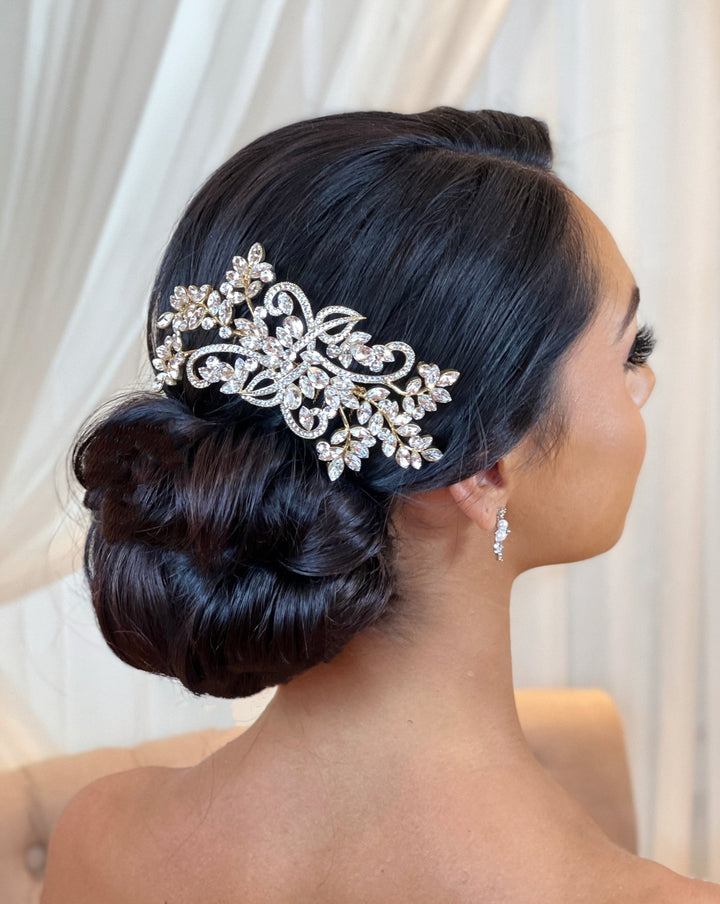 A woman with dark hair styled in a low bun is adorned with the Flowing Crystal Branch Comb style no. 242322 by Swarovski. She wears small, elegant earrings, and the softly blurred background enhances the focus on her sparkling hairstyle.