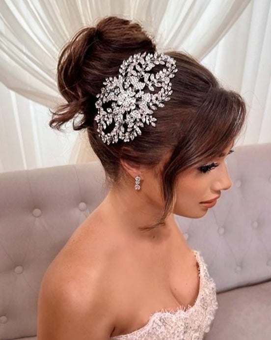 A woman in a strapless white gown, her elegant updo adorned with the intricate floral design of Crystal Bloom Hair Comb style no. 242717, sits on a light grey tufted sofa with sheer white curtains behind her.