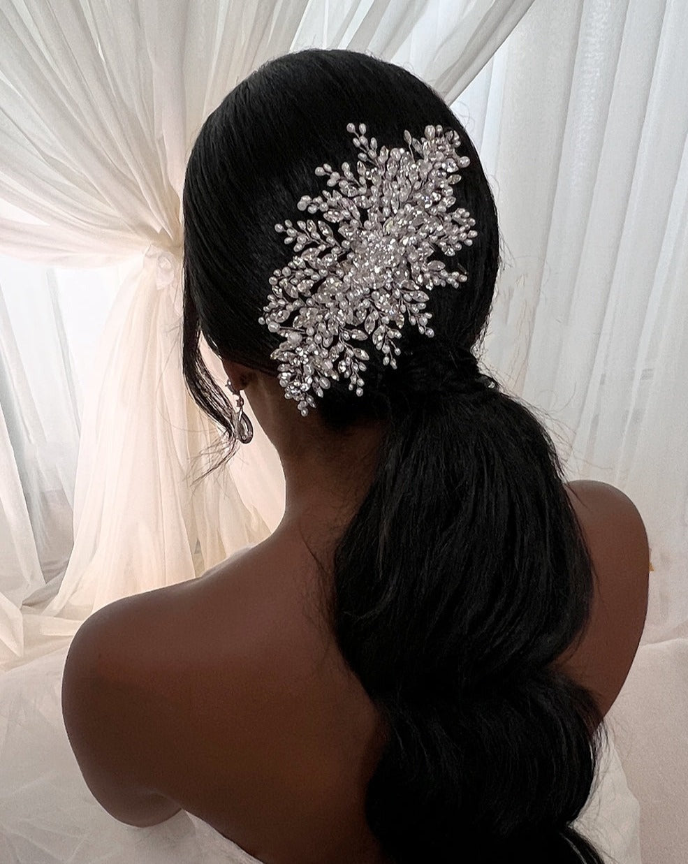 Rear view of a person with long, dark hair in a low ponytail adorned with the Classic Swarovski Crystal Bridal Comb (style no. 242646) resembling a snowflake. They are wearing CZ wedding earrings and sitting in front of flowing white drapery.