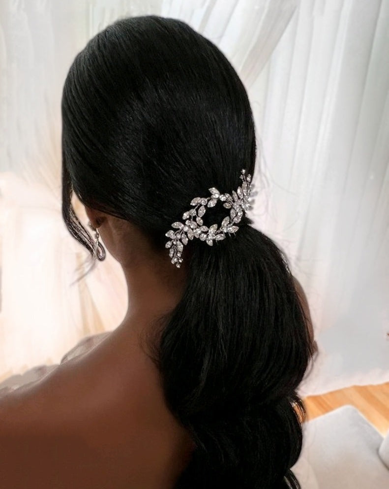 With long, dark hair tied in a low ponytail, a woman adorns herself with the Dainty Swarovski Crystal Bridal Comb style no. 242615. She complements it with subtle earrings, sitting gracefully against sheer white curtains.