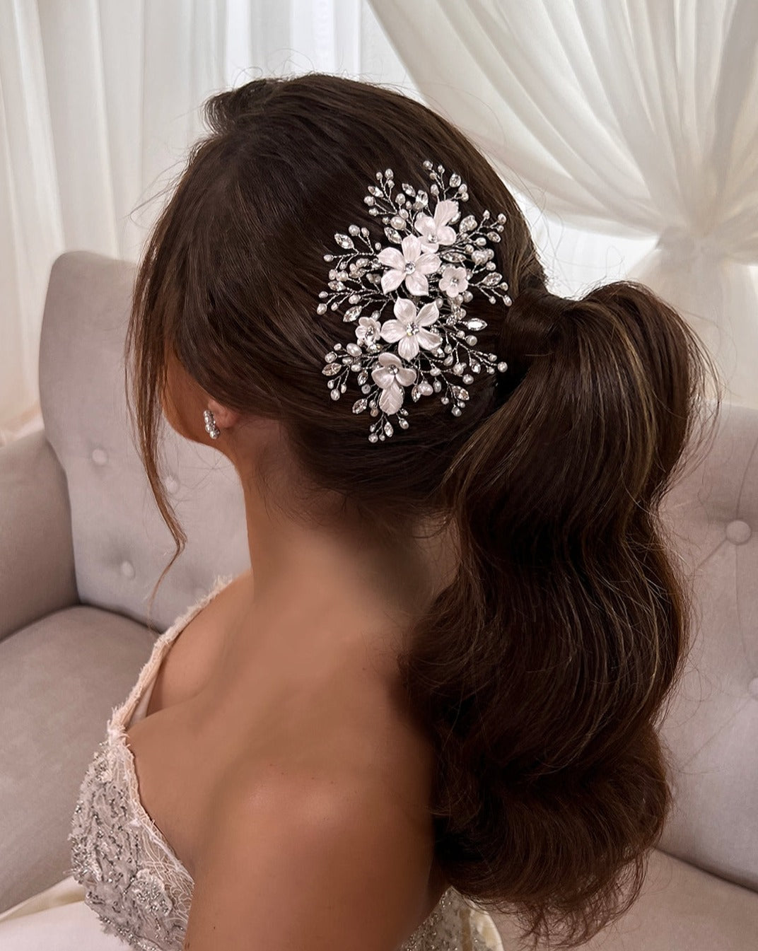 A woman with long, wavy brown hair in a ponytail sits on a beige chair. Her hair is decorated with the Porcelain Flower Bouquet Swarovski Crystal Hair Comb (style no. 242722) featuring floral and pearl details. She wears a strapless lace wedding dress and small, sparkling earrings against a white curtain background.