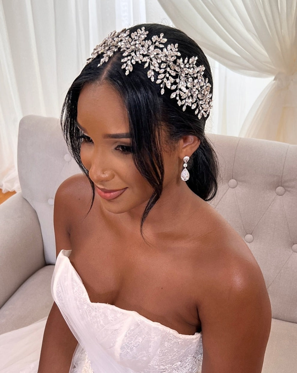 A bride sits on a beige sofa in a white strapless gown. Her hair is styled with the Glistening Cluster Swarovski Crystal Vine, style no. 3951567, featuring leaves and flowers. She wears pearl drop CZ bridal earrings and smiles softly, with sheer white curtains in the background.