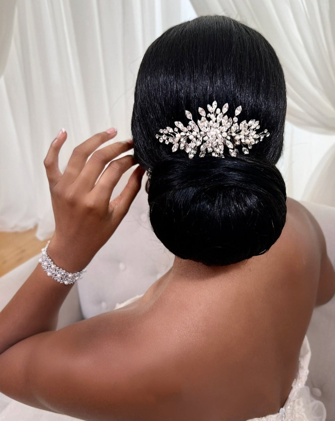 A woman with dark hair in a bun adjusts her hair with a left hand, while wearing the Dainty Swarovski Crystal Hair Comb style no. 242731 by Swarovski and an ornate bracelet. The background features soft white drapery.