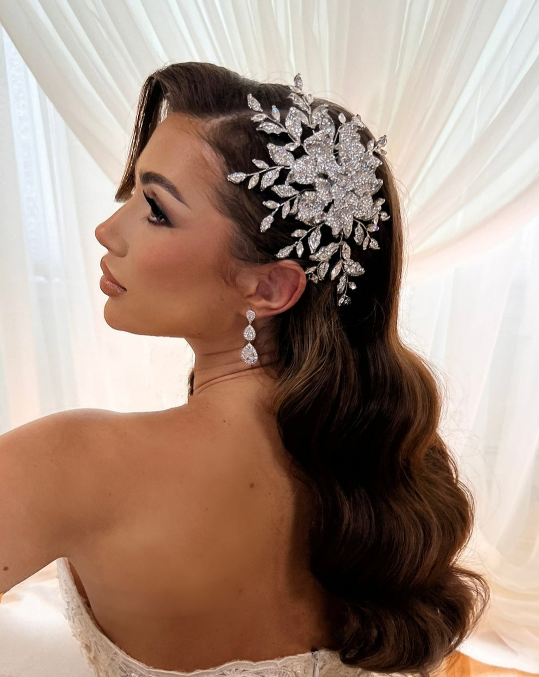 A woman with long brown hair, adorned with an Enchanting Crystal Rose Bridal Hair Comb (style no. 242587), stands before flowing white curtains in a strapless white dress and dangling earrings, looking over her shoulder. The atmosphere exudes elegance and bridal charm.