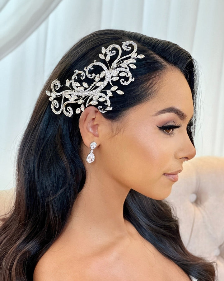 A woman with long, dark hair wears a Swarovski Crystal Swirl Bridal Comb (style no. 242370), its intricate swirls and leaf designs call to mind a spray of flowers. Her elegant, dangling earrings are highlighted against the softly blurred background, emphasizing her profile and accessories.