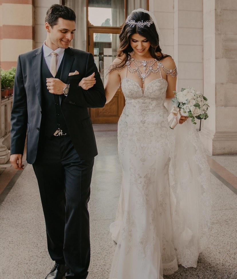 A bride and groom walk arm in arm. the bride wears a sparkling fit and flare gown with shoulder jewelry and her hair down with a crystal hair vine