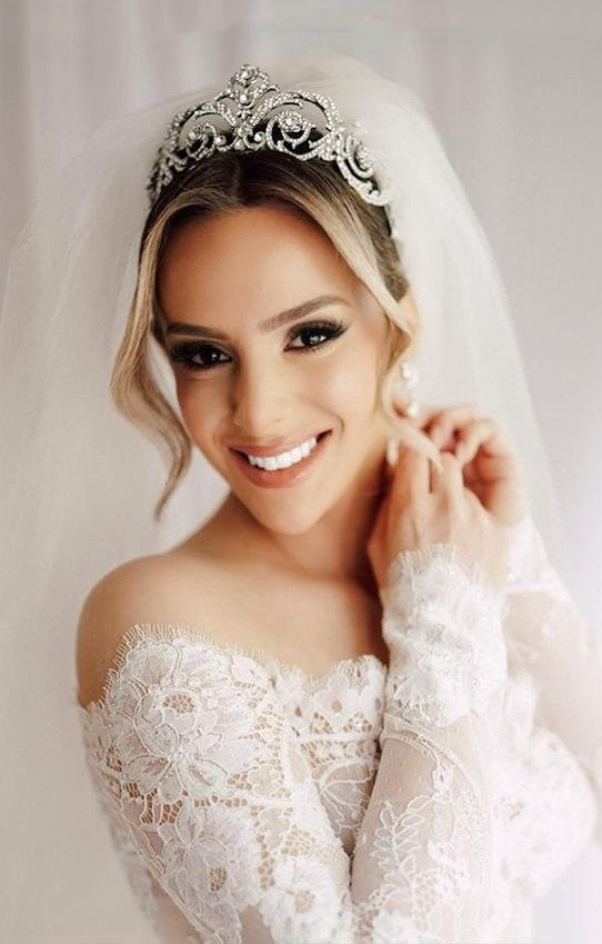 Bride wearing an off-the-shoulder lace wedding gown, crystal tiara, and veil, smiling as she adjusts her earring.