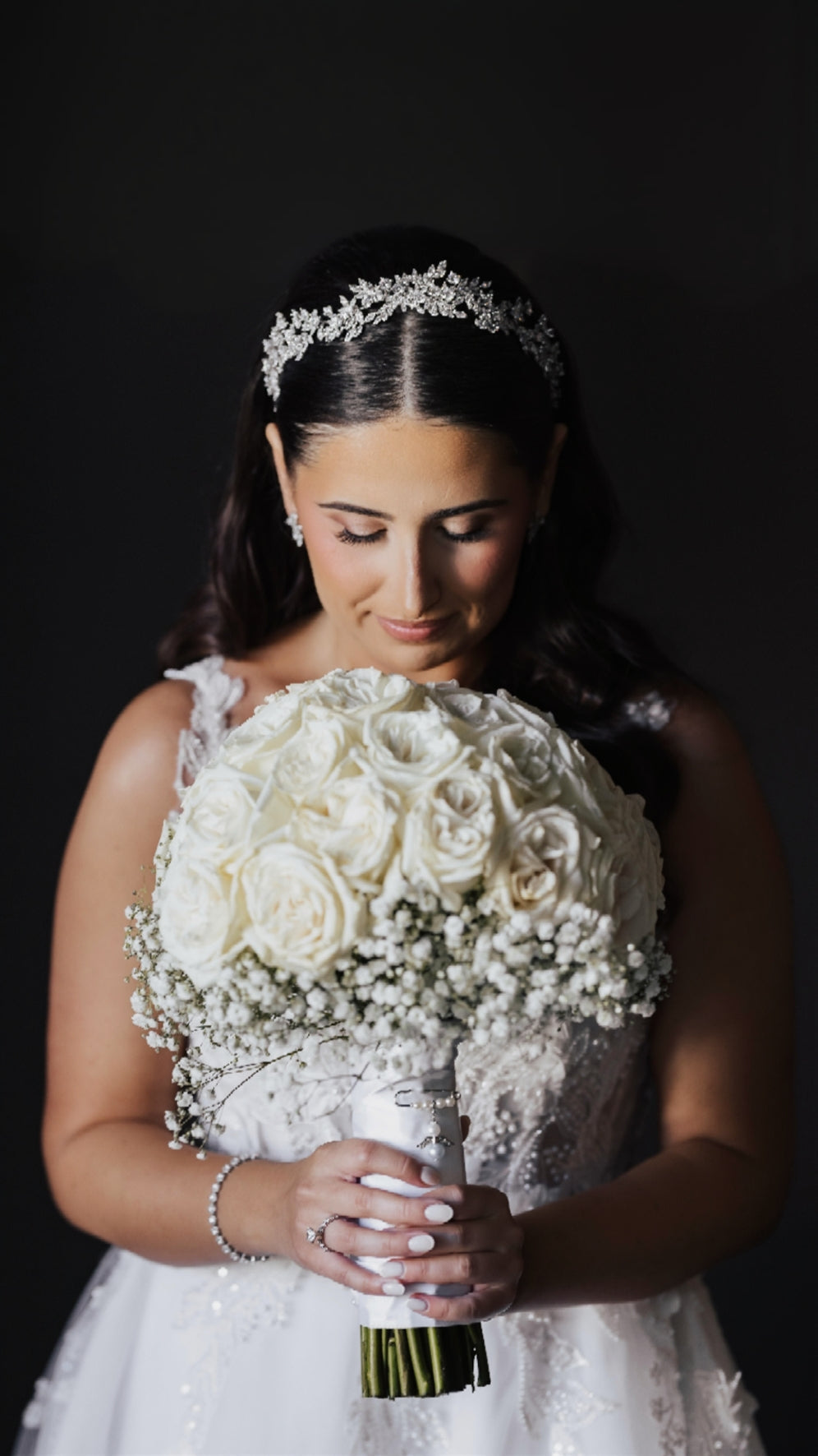 Bride Grazia looking down at white rose and baby's breath bouquet wearing crystal bridal headpiece and bracelet from Bridal Styles Boutique