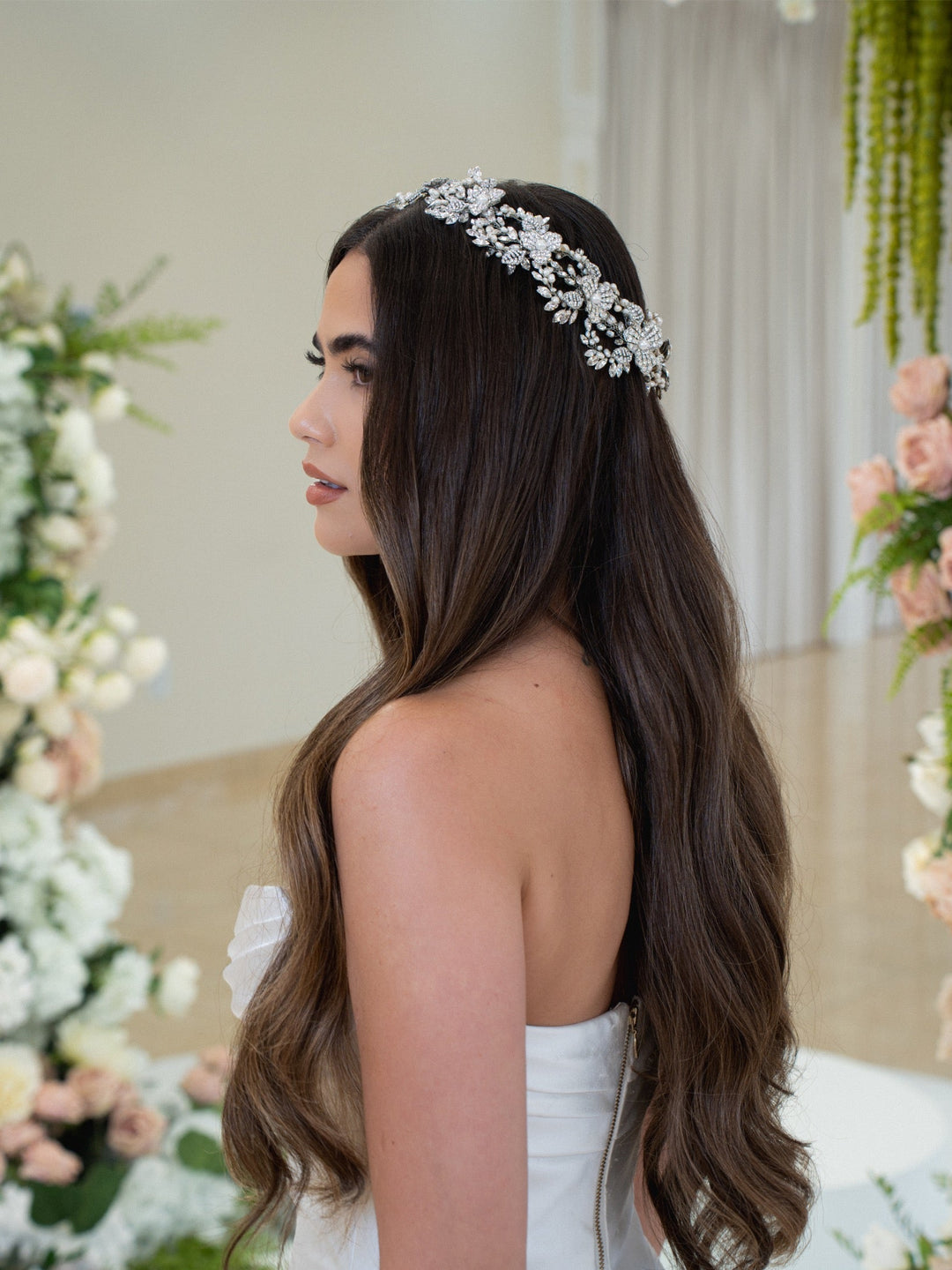 A bride with long dark wavy hair wearing a crystal and pearl floral bridal halo headband, seen from a three-quarter back angle in a strapless white wedding gown, surrounded by white and blush floral arrangements