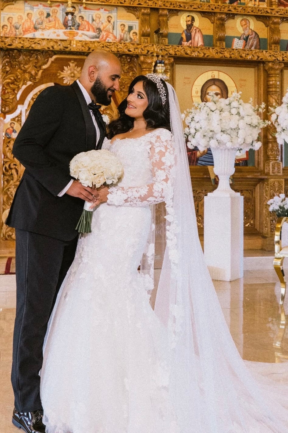 Bride Jenna in a custom crystal tiara headband, lace long-sleeve mermaid gown, and lace-edged cathedral veil, smiling at her groom at the church altar