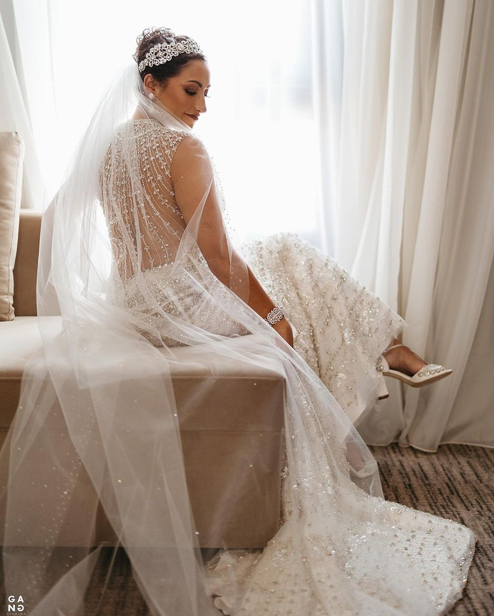 Bride in ornate beaded cathedral wedding veil and embellished lace gown sitting on bench