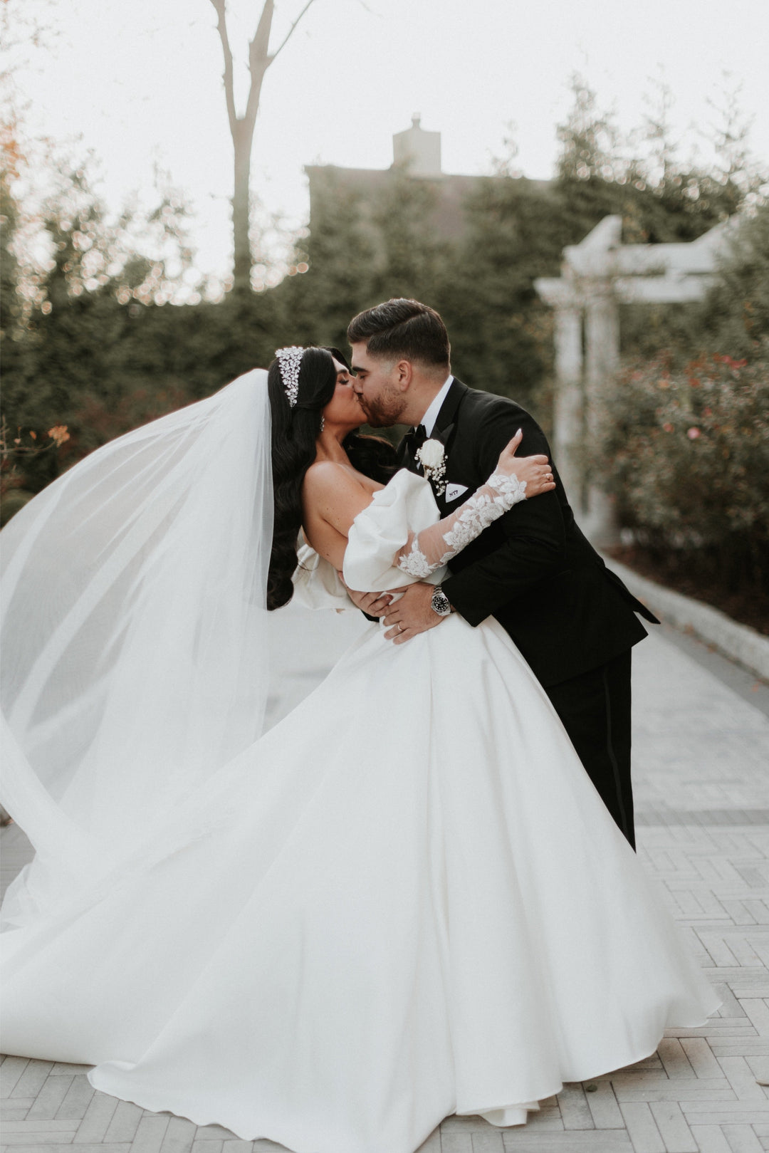 Maria kissing groom outdoors at Florentine Gardens, crystal crown and lace royal cathedral veil billowing dramatically, wearing Pnina Tornai ball gown with lace gauntlets