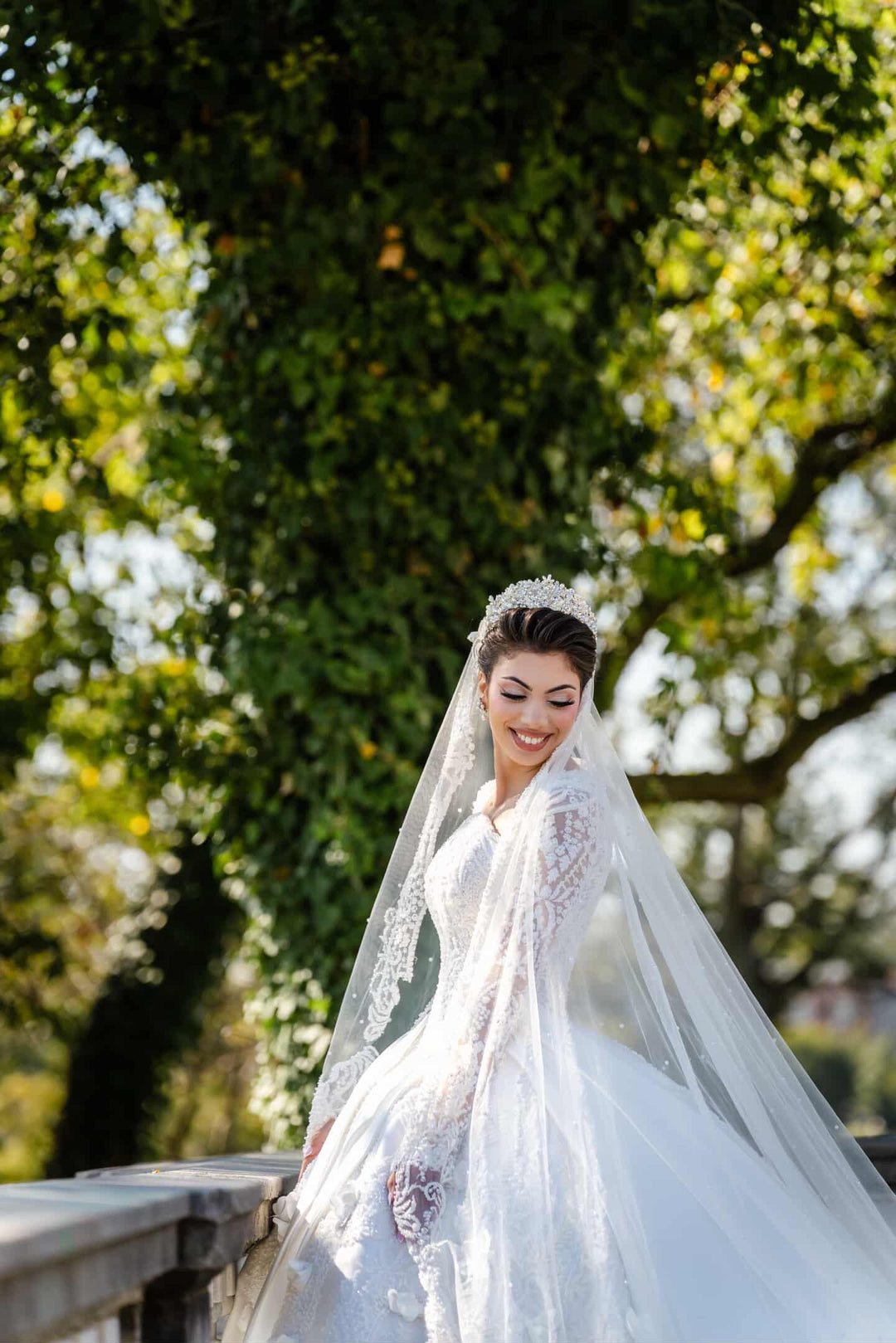 Real bride Moufideh wearing a pearl and Swarovski crystal bridal crown with matching crystal earrings from Bridal Styles Boutique, paired with a heavily embellished lace ball gown and cathedral veil, smiling outdoors in natural light.