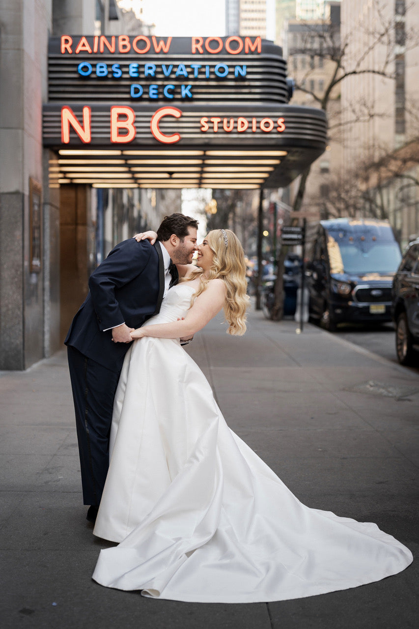 A bride and groom kiss in front of the Rainbow Room at Dusk. The bride wears a ball gown, her long curly hair down, and a crystal wedding hair vine 