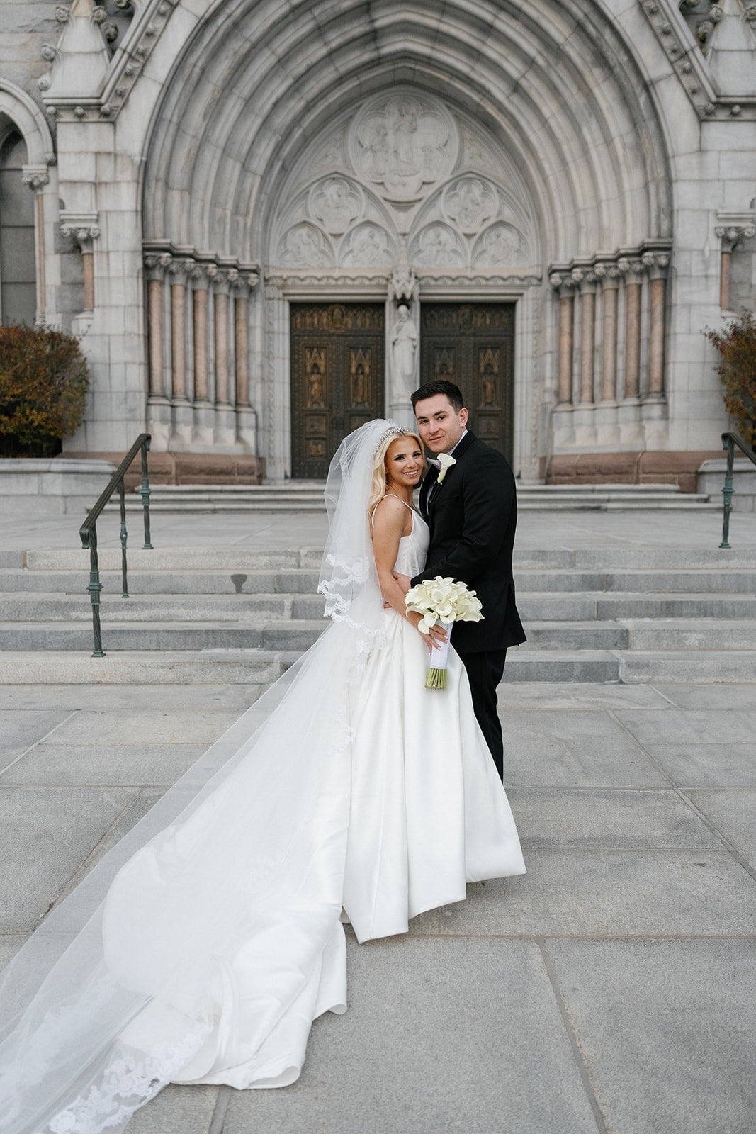Bride Samantha and her groom posed on the steps of a grand stone cathedral, her lace-edged royal cathedral veil trailing behind her ballgown; she wears a Swarovski crystal headband and carries a white calla lily bouquet
