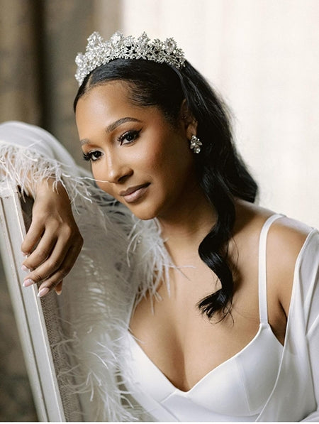 A bride in a white robe wears a regal bridal crown and jeweled earrings, leaning gracefully against a surface.