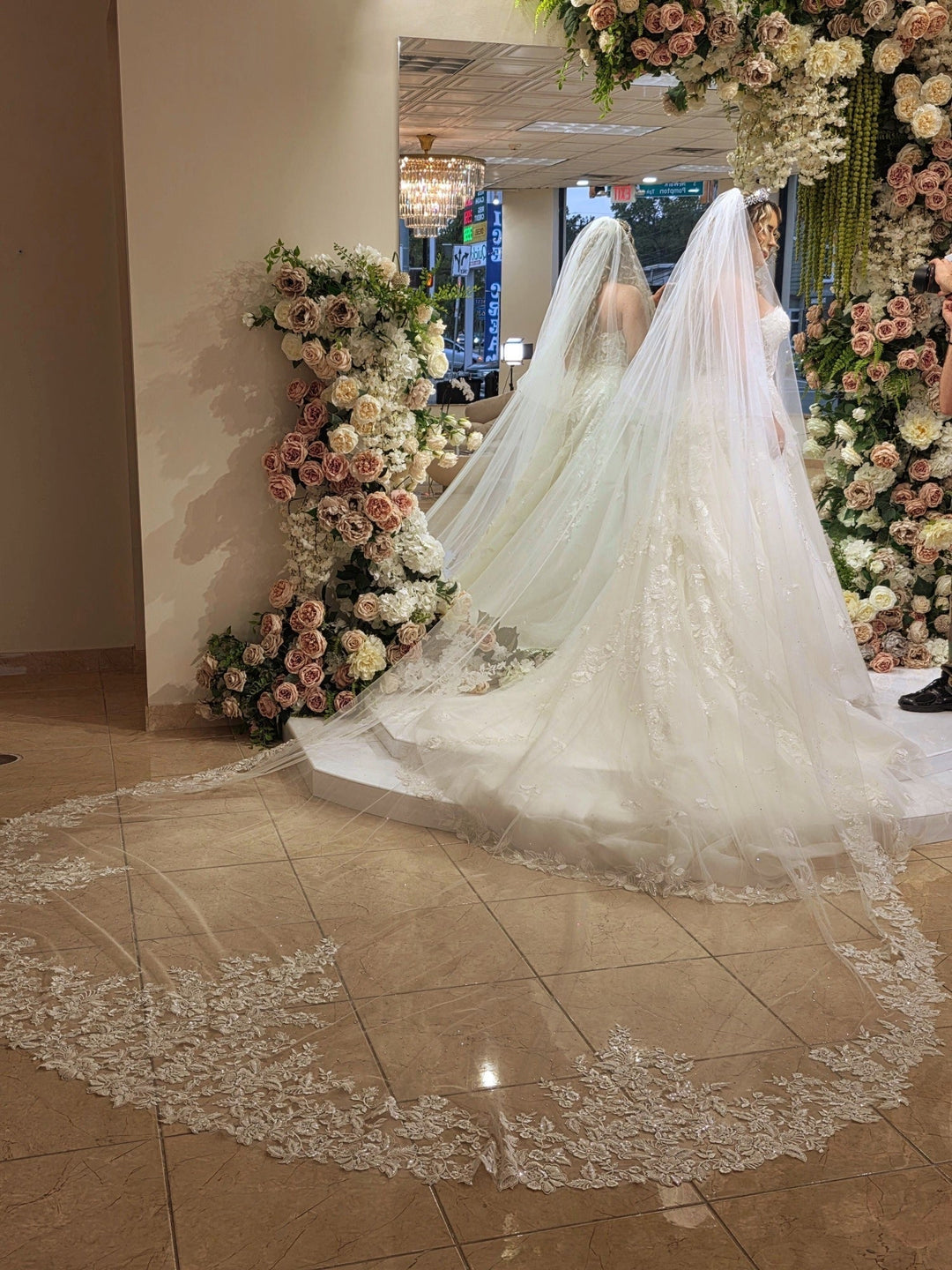 Bride's long, lace-edged wedding veil trailing across the floor amid blush and ivory rose arrangements before her ceremony.