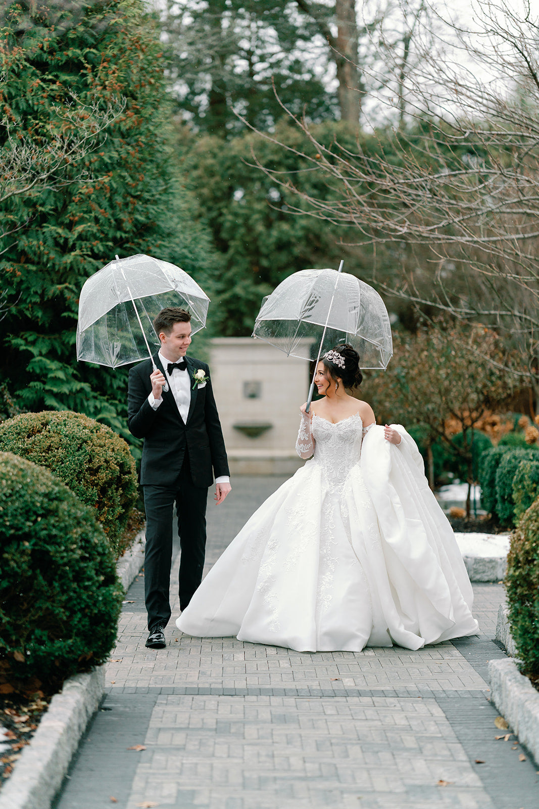 Bride Alexa and groom Liam walking together under clear umbrellas, bride wearing custom pearl and crystal headpiece.