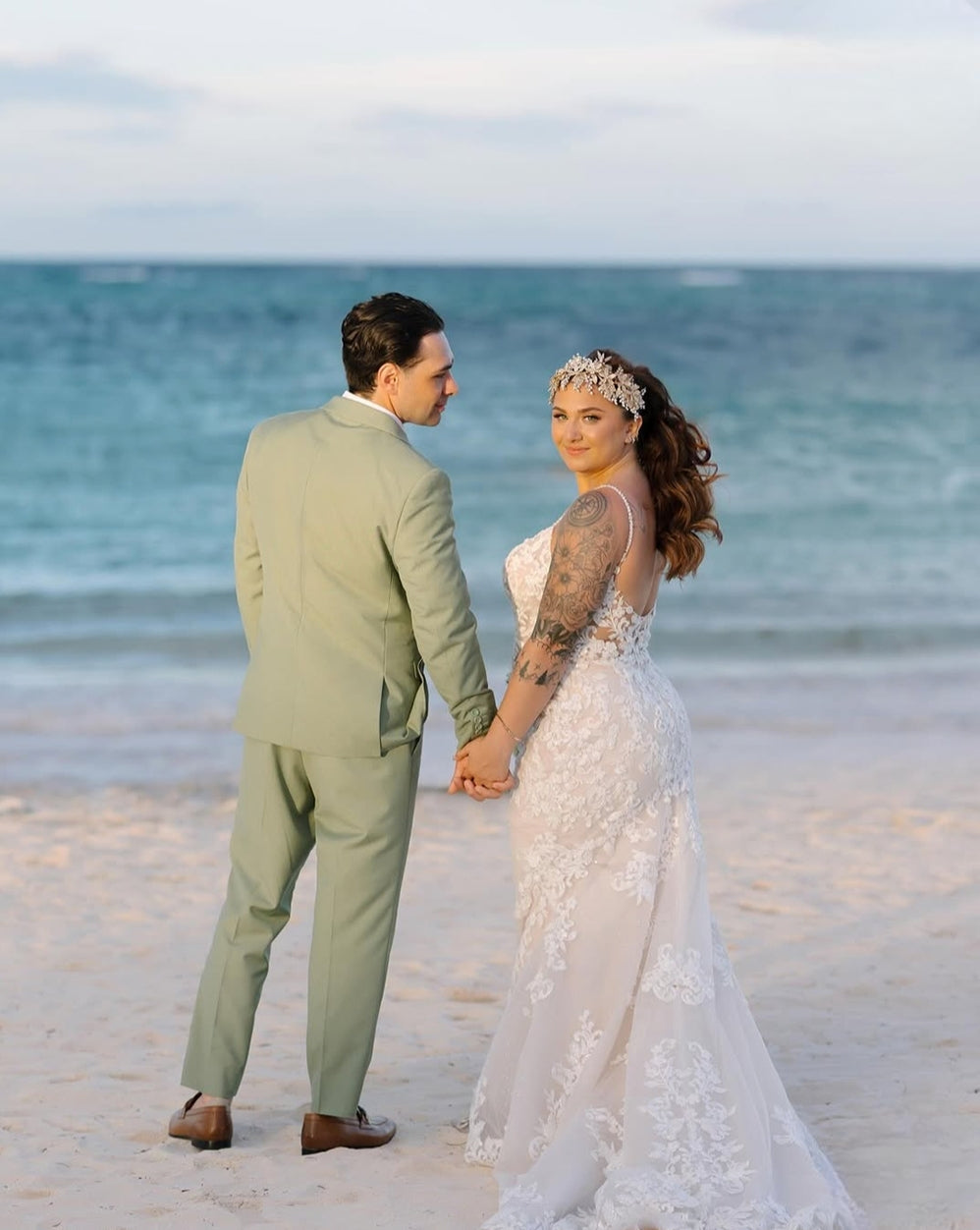 Bride Ariana and her groom holding hands on the beach in Punta Cana, bride wearing a custom silver and gold leaf crown headpiece with her lace wedding gown