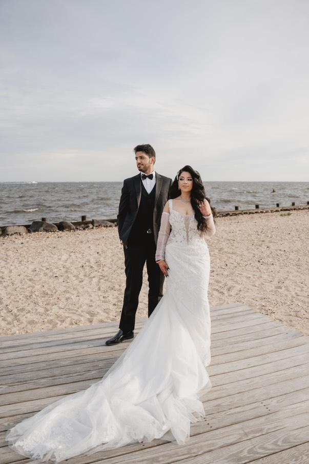 Bride Grace stands beside her groom on a beach boardwalk, wearing a lace wedding gown and custom Swarovski crystal hair vine from Bridal Styles Boutique.