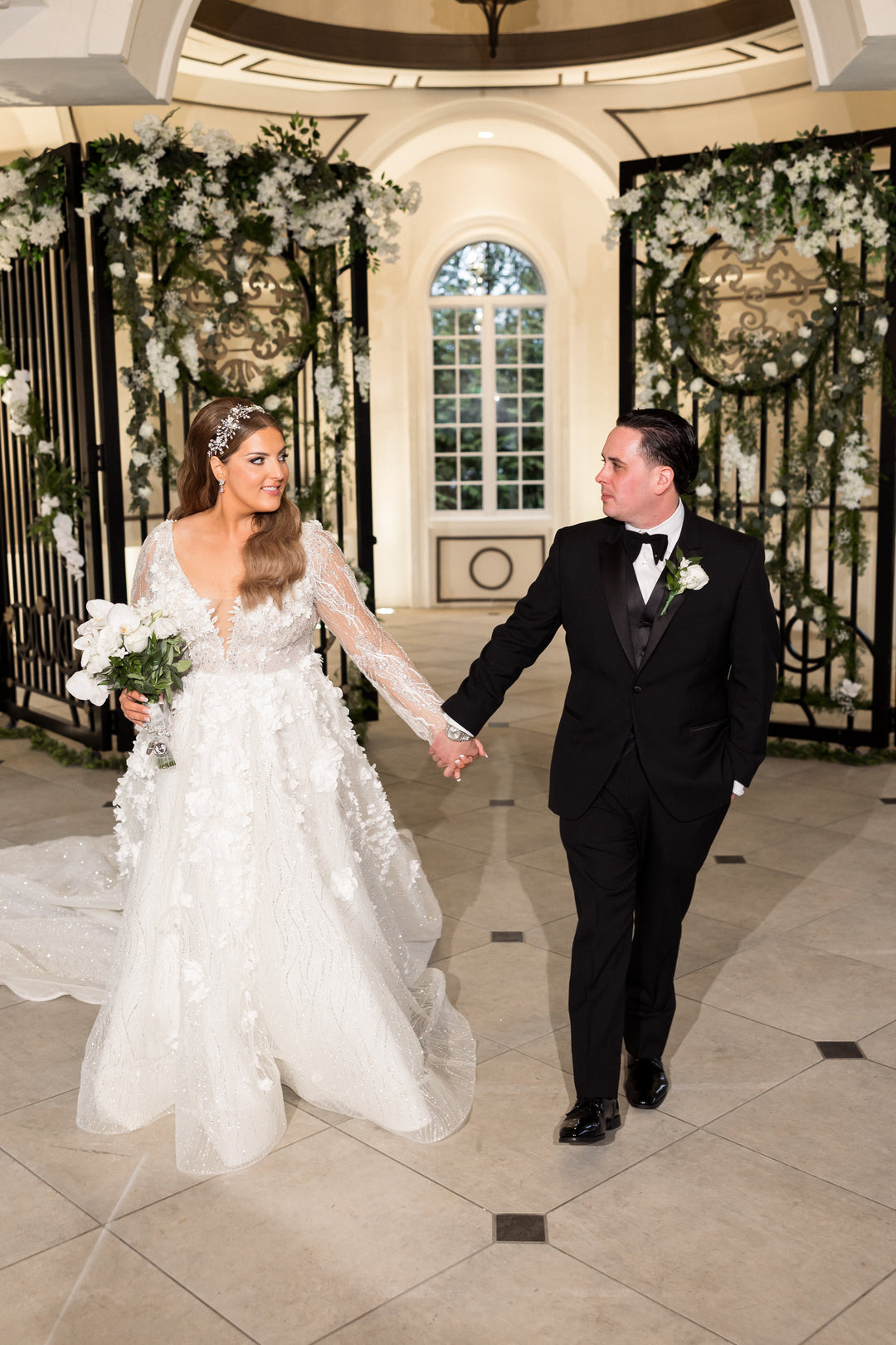 Bridal Styles bride Briana wearing a lace wedding gown and crystal hair vine with porcelain flowers, walking hand-in-hand with her groom beneath a floral arch