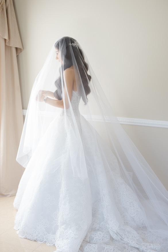 Bride in a strapless lace gown and custom cathedral veil, wearing a Swarovski crystal headpiece, standing by a window in soft natural light.