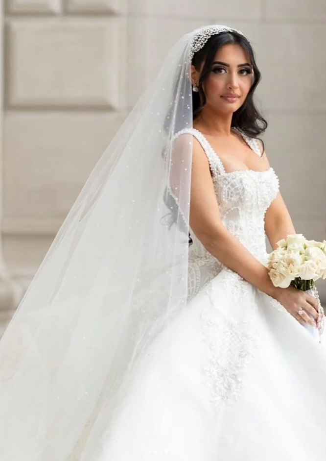 Bride with long dark hair wears a pearl-studded headband and cathedral veil over a beaded ballgown wedding dress, holding a bouquet of cream roses.