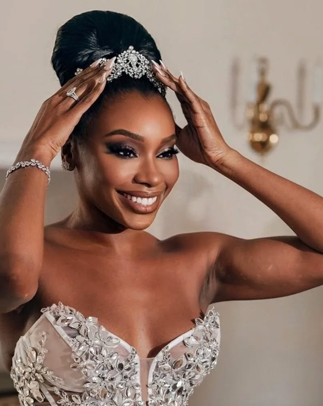 Smiling bride wearing a crystal tiara with a sleek high bun and strapless floral-appliqué gown, captured mid-moment adjusting her headpiece