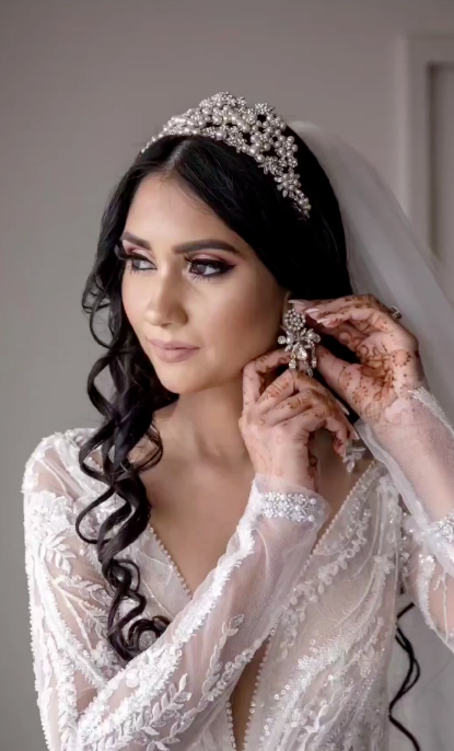 Bride putting on chandelier statement wedding earrings while wearing ornate crystal tiara, demonstrating maximalist bridal styling
