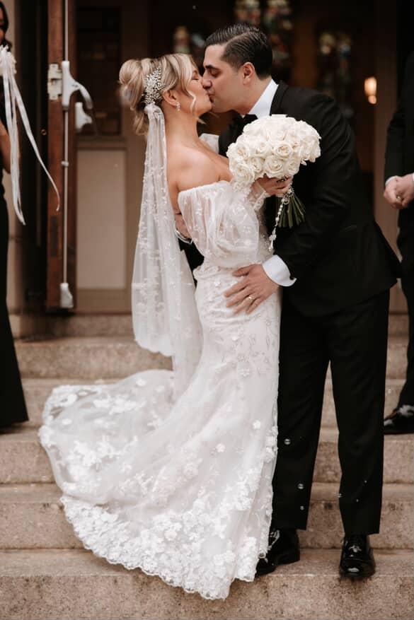 Bride Elyse in off-shoulder lace wedding gown with custom floral crystal headpiece and cathedral veil kissing groom on church steps holding white rose bouquet
