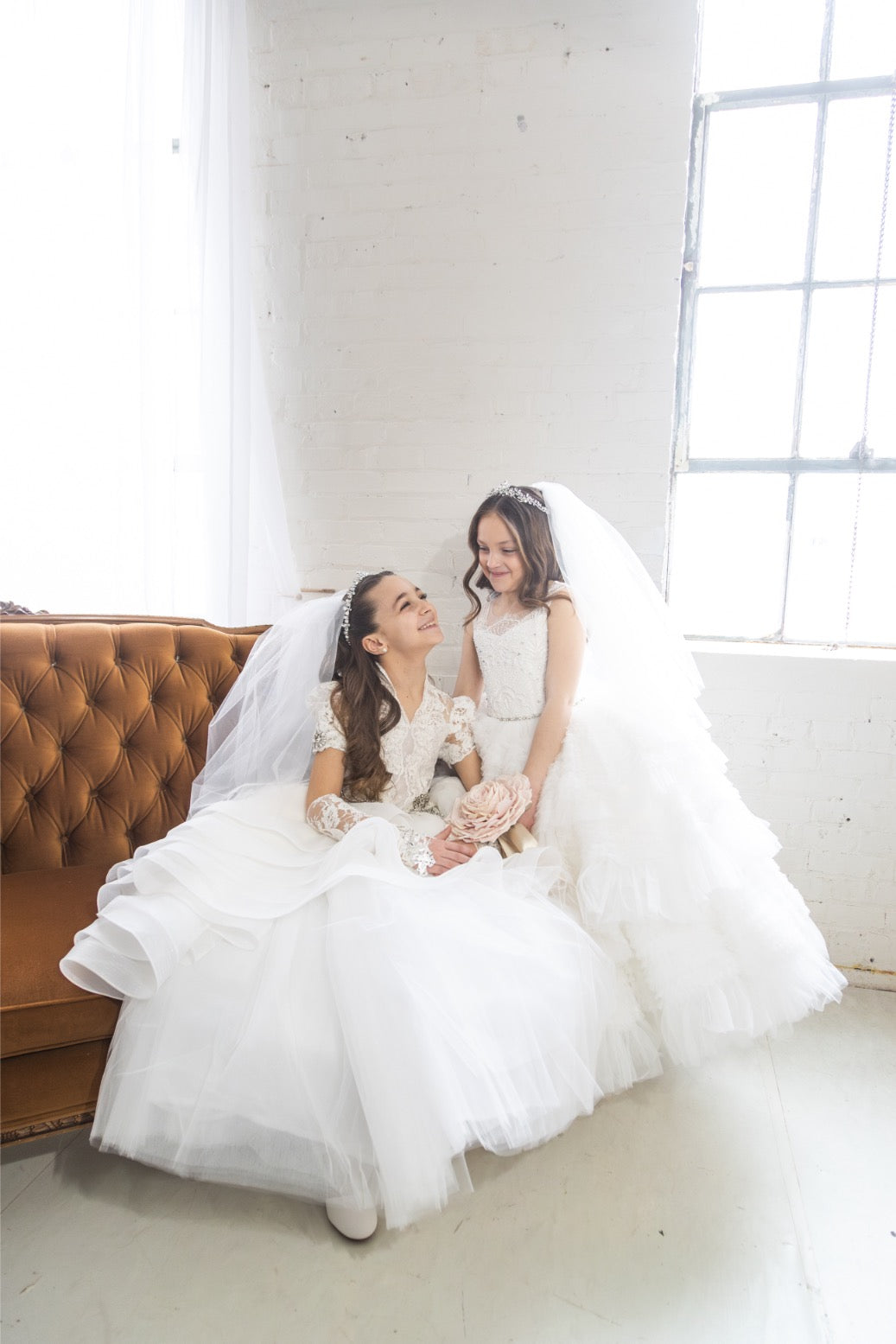 Two girls in First Communion dresses wearing crystal tiaras and veils, seated together on a velvet sofa