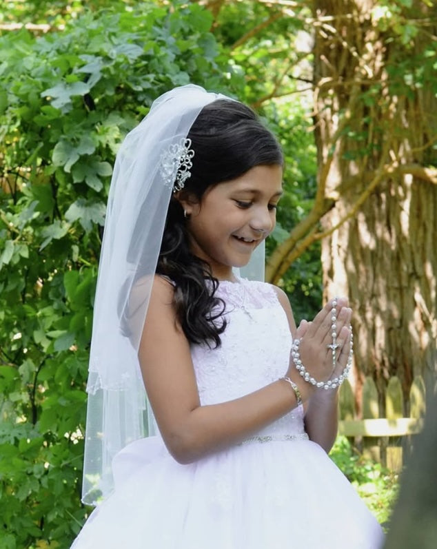 a little girl on her first communion, her hands together in prayer with a rosary. She is wearing a crystal comb and communion veil