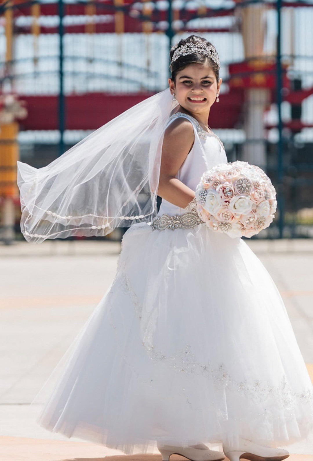 Isabella smiling at Coney Island in her first communion dress, wearing her mother Tina's crystal hair vine and drop earrings, veil catching the wind