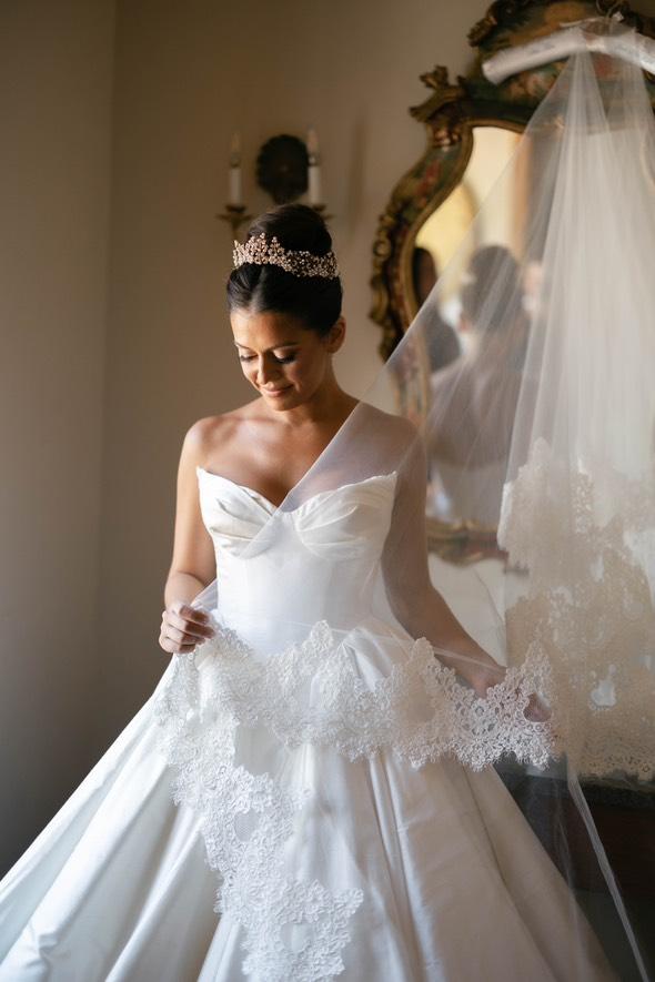 Jacqueline stands in soft light holding her cathedral veil, her pearl and crystal tiara and structured ballgown creating a timeless bridal look.