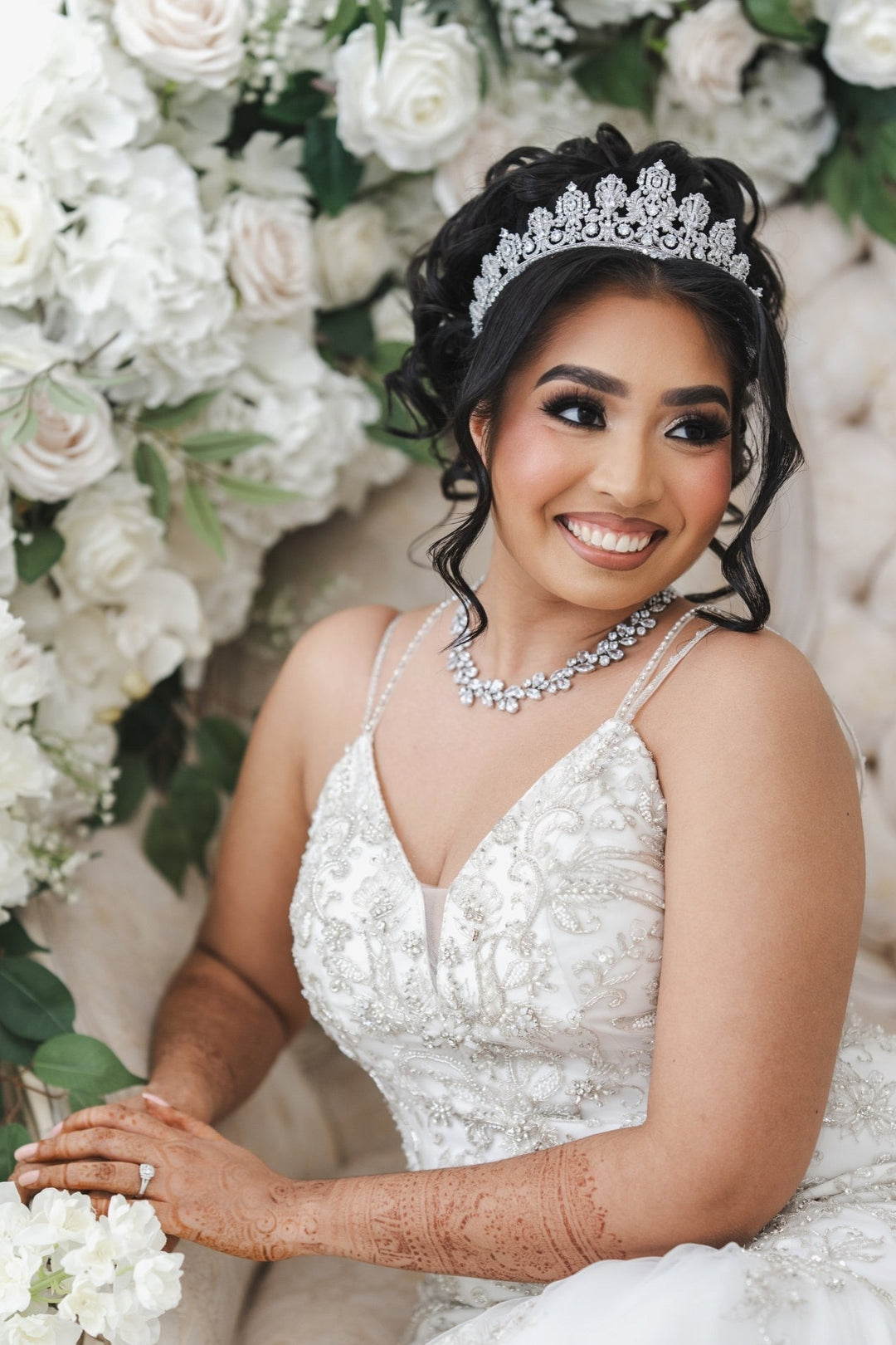 Mary Ann smiling in a beaded wedding gown, crystal tiara, and layered crystal necklace, seated in front of a white floral wall with henna on her arms and hands.