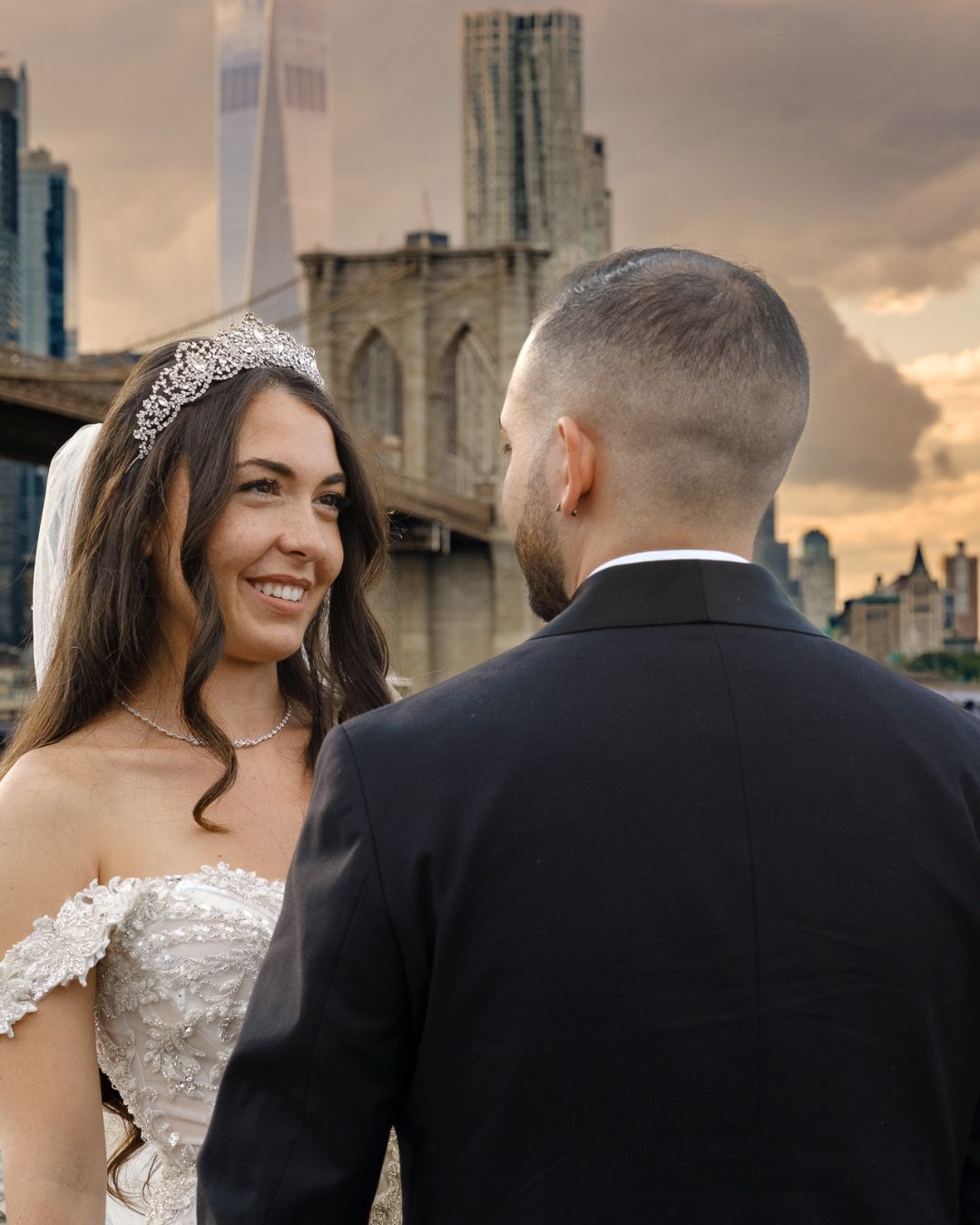 Bride Melissa in a crystal crown and off-the-shoulder gown smiling at her groom in front of the Brooklyn Bridge at sunset