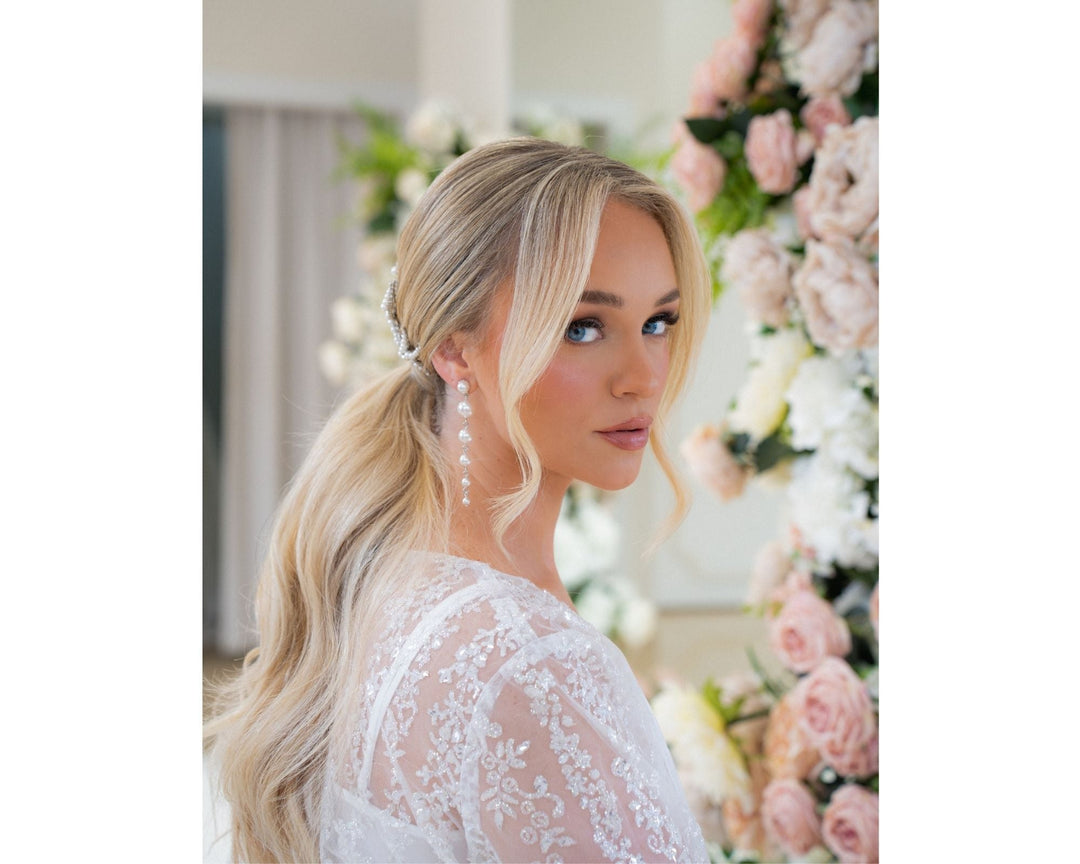 Blonde bride wearing long pearl dangle bridal earrings with a low ponytail, glancing over her shoulder near blush garden roses