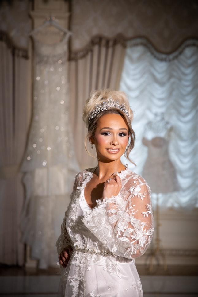 Bride Claudia wearing her Swarovski crystal bridal tiara and lace robe, glowing as she gets ready with her wedding gowns in the background.