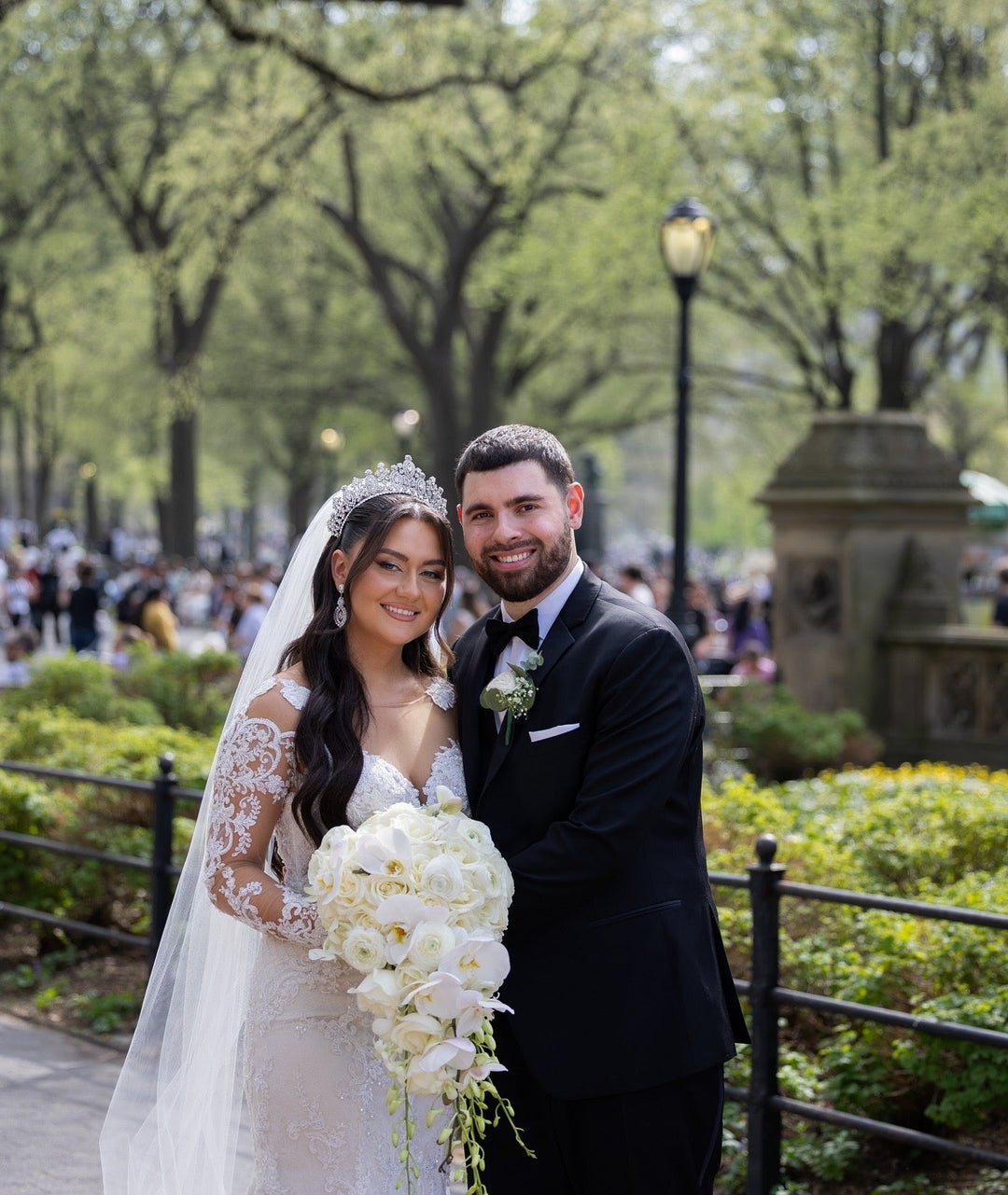  real bride Eldina in a lace wedding gown with a crystal crown, matching earrings, and cascading white rose and orchid bouquet, smiling with her groom in a garden setting.