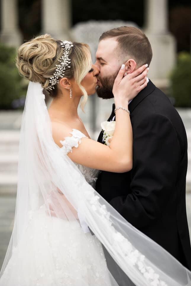 Bride Rebecca kisses her groom while wearing a crystal wedding halo, lace veil, and off-the-shoulder gown.