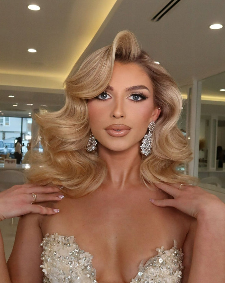 A woman with voluminous, wavy blonde hair and a strapless embellished gown poses with her hands near her collarbone in an elegant room, wearing Studded Pearl and Crystal Chandelier Earrings (style no. E218).